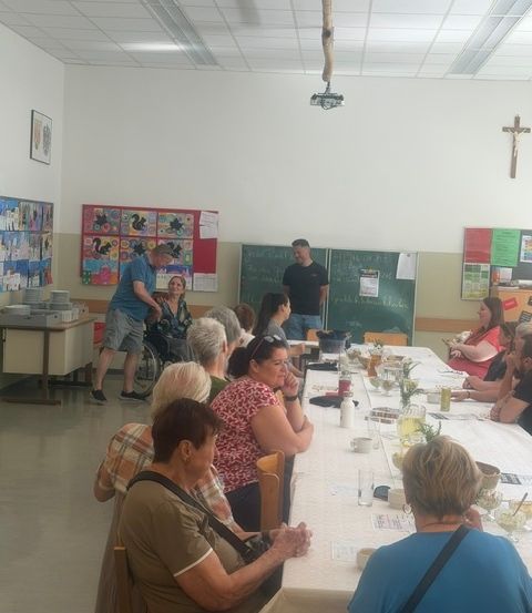 Eine Gruppe von Menschen, darunter eine ältere Frau im Rollstuhl, sitzt um einen langen Tisch in einem Raum. Ein Mann steht in der Nähe des Tisches und ein anderer Mann steht in der Nähe einer Tafel. Auf dem Tisch befinden sich verschiedene Gegenstände und ein Kreuz ist an der Wand montiert.