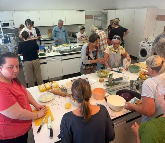 Eine Gruppe von Menschen ist in einer Küche und bereitet Essen zu. Einige stehen und andere sitzen. Auf dem Tresen befinden sich verschiedene Zutaten und Utensilien.