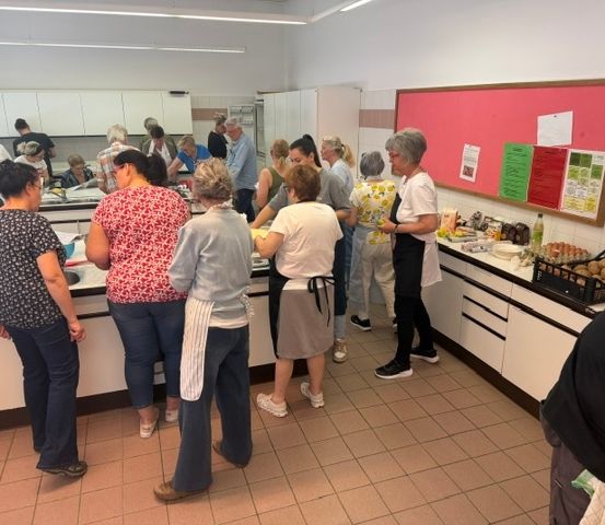 Eine Gruppe von Menschen in einer Küche, einige mit Schürzen, andere ohne. Sie stehen um einen Tresen mit Essen und Zutaten herum.