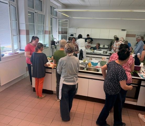 Eine Gruppe von Menschen in einer Küche, einige stehen, einige sitzen, mit einer Frau in einem roten Shirt und orangefarbenen Hosen, die hervorsticht.