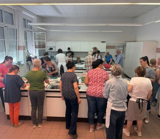 Eine Gruppe von Frauen in einer Küche, einige stehend und andere sitzend, beschäftigt mit der Zubereitung von Speisen, mit einer Mikrowelle und einem Kühlschrank im Hintergrund.