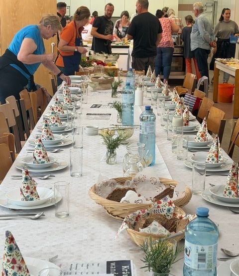 Ein langer Tisch, der für eine Mahlzeit mit Blumenservietten, Wasserflaschen und verschiedenen Gerichten gedeckt ist. Im Hintergrund stehen Menschen und bereiten Essen zu.