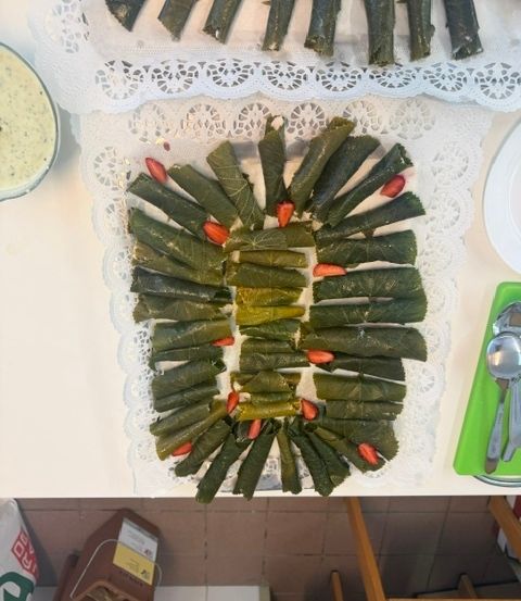 Ein Tisch zeigt eine Platte mit grünen Blattrollen mit roten Paprikaschoten, die in einem kreisförmigen Muster angeordnet sind. Eine weiße Spitzendeckchen und eine weiße Schüssel mit grüner Sauce sind ebenfalls sichtbar.