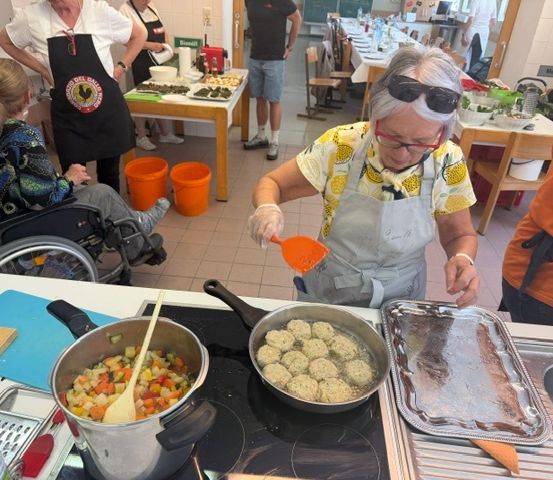 Eine ältere Frau mit Brille und Schürze kocht in einer Küche mit anderen Menschen. Sie benutzt einen Schöpflöffel, um einen Topf mit Essen auf dem Herd zu rühren.