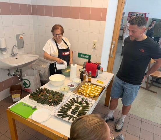 Eine Frau in einem weißen Shirt und einer Schürze bereitet Essen auf einem Tisch zu, während ein Mann in der Nähe steht. Der Tisch ist mit verschiedenen Gerichten gefüllt, und ein Waschbecken ist im Hintergrund zu sehen.