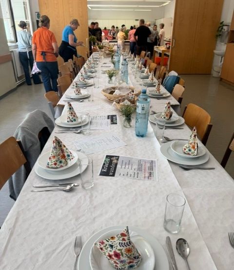 Ein langer Esstisch, der für eine Mahlzeit mit Blumenservietten, Tellern, Gläsern und Besteck gedeckt ist. Im Hintergrund stehen und unterhalten sich Menschen.