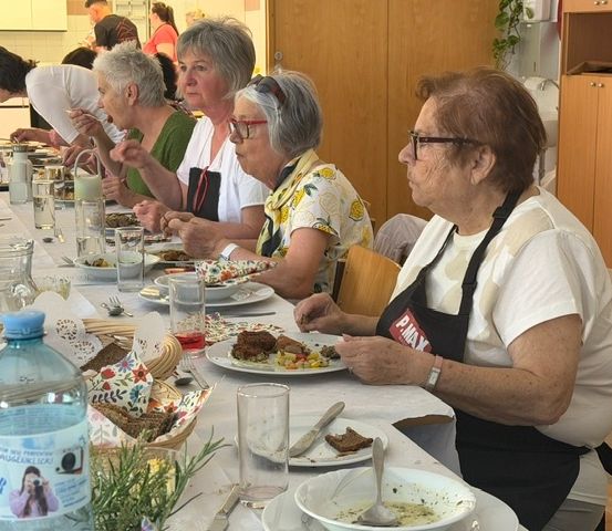 Eine Gruppe älterer Frauen sitzt an einem langen Tisch, isst und plaudert. Sie tragen Schürzen und haben Teller mit Essen vor sich.