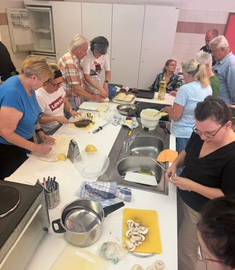 Eine Gruppe von Menschen versammelt sich um einen Küchentresen und bereitet Essen zu. Sie verwenden verschiedene Küchenutensilien und Zutaten, einschließlich eines Schneidebretts mit Pilzen, einer Schüssel und einem Topf. Die Küche hat weiße Schränke und geflieste Wände.