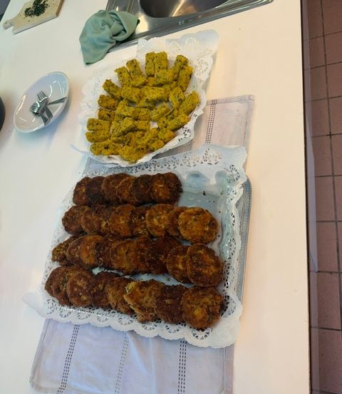 Zwei Tabletts mit Essen, eines mit gelben Pasteten und das andere mit braunen Pasteten, sind auf einer weißen Arbeitsplatte ausgestellt. Ein Messer und ein Schneidebrett sind auf der Arbeitsplatte platziert. Ein Waschbecken mit einem Wasserhahn befindet sich auf der rechten Seite. Ein weißes Tuch ist auf der Arbeitsplatte platziert.