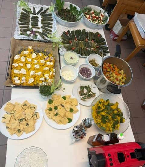 Ein Tisch ist mit verschiedenen Gerichten gefüllt, darunter gelbe Quadrate, grünes Gemüse und eine große Schüssel mit gemischtem Gemüse. Schüsseln, Teller und ein Topf sind ebenfalls vorhanden.