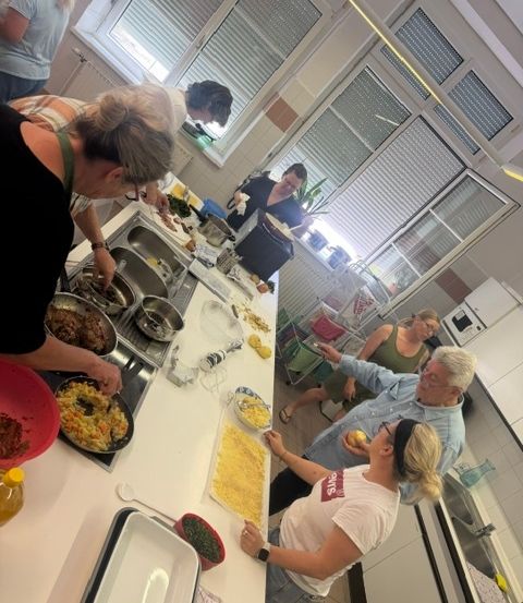 Eine Gruppe von Menschen bereitet in einer Küche Essen zu. Sie stehen um einen langen Tisch mit verschiedenen Gerichten und Utensilien herum. Einige von ihnen tragen Brillen und Armbänder. Die Küche hat einen gefliesten Boden, ein Fenster mit Jalousien und eine Glastür. Es gibt auch einige Topfpflanzen und eine Mikrowelle auf der Theke.
