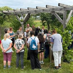 Eine Gruppe von Menschen steht in einem Garten, einige hören einem Mann zu, der unter einem hölzernen Pergola spricht.
