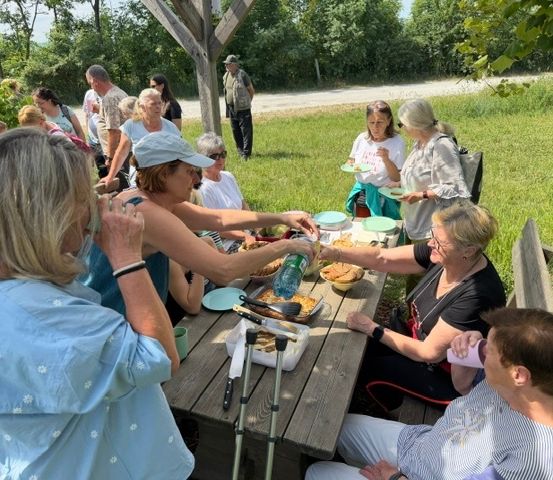 Eine Gruppe von Menschen, hauptsächlich Frauen, versammelt sich um einen Picknicktisch auf einer Wiese. Einige sitzen, andere stehen und alle unterhalten sich. Der Tisch ist mit Tellern, Schüsseln und Besteck beladen. Im Hintergrund steht ein Mann in der Nähe eines Baumes.
