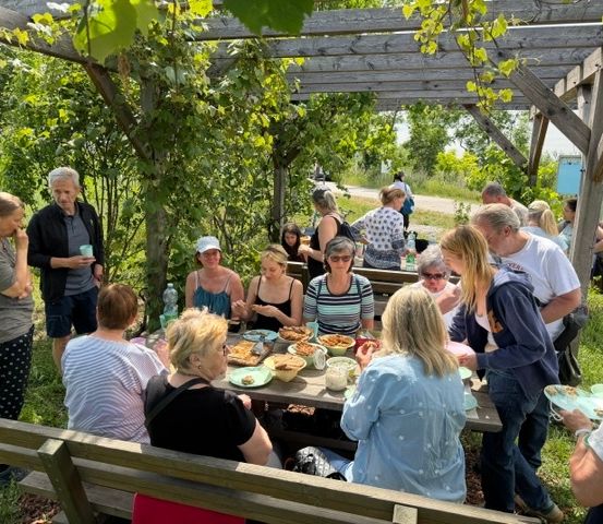 Eine Gruppe von Menschen versammelt sich um einen Tisch im Garten und genießt eine Mahlzeit. Der Tisch ist mit Essen beladen, und die Gruppe besteht aus Männern und Frauen. Einige sitzen auf Bänken, während andere stehen. Die Umgebung ist im Freien, mit viel Grün und einer hölzernen Struktur im Hintergrund.
