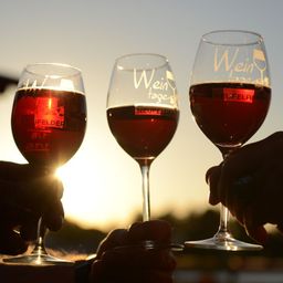 Drei Weingläser mit rotem Wein werden vor einem Sonnenuntergangshintergrund von Händen gehalten. Die Gläser haben die Worte 'Wein' und 'Wein tage' darauf.