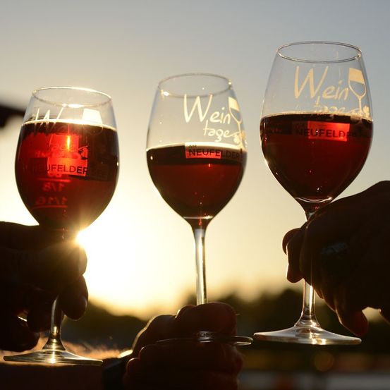 Drei Weingläser mit rotem Wein werden vor einem Sonnenuntergangshintergrund von Händen gehalten. Die Gläser haben die Worte 'Wein' und 'Wein tage' darauf.