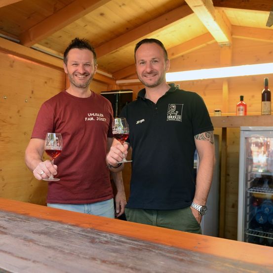 Zwei Männer stehen hinter einer Theke, lächeln und halten Weingläser. Einer trägt ein weinrotes T-Shirt, der andere ein schwarzes Poloshirt. Hinter ihnen Holzwände und ein Kühlschrank mit Flaschen.