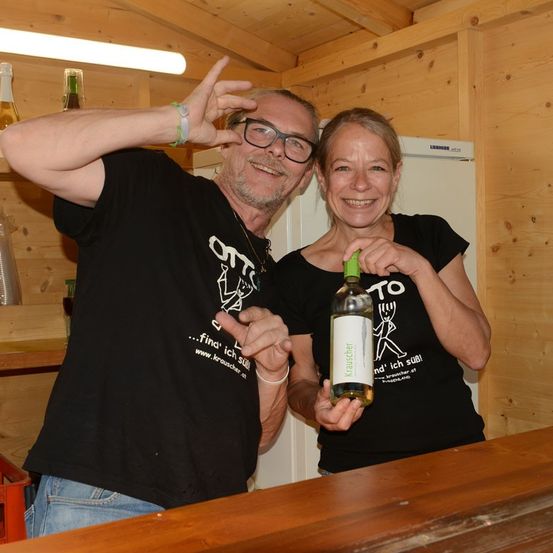 Ein Mann und eine Frau in passenden schwarzen T-Shirts posieren mit einer Flasche Wein in einem hölzernen Raum.
