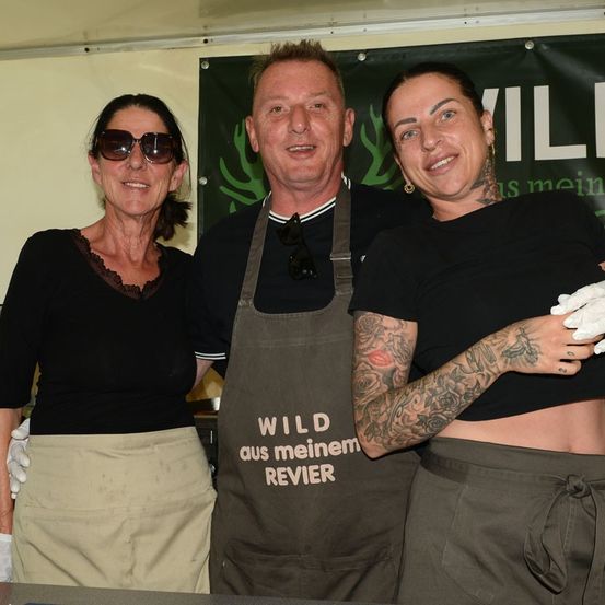 Drei Personen stehen in einer Küche, tragen Schürzen. Der Mann hat eine Kamera um den Hals, und die Frauen tragen weiße Handschuhe. Das Banner hinter ihnen lautet 'Wild aus meinem Revier'.