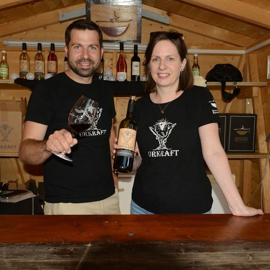 Ein Mann und eine Frau in schwarzen Urkraft-T-Shirts posieren mit einer Flasche und einem Glas Wein in einer rustikalen Umgebung mit Holzregalen und Flaschen im Hintergrund.