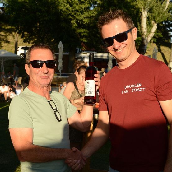 Zwei Männer lächeln und schütteln sich die Hände in einer Außenumgebung. Ein Mann hält eine Flasche Wein. Hinter ihnen sitzen Leute unter Sonnenschirmen. Bäume und ein weißes Auto sind im Hintergrund.