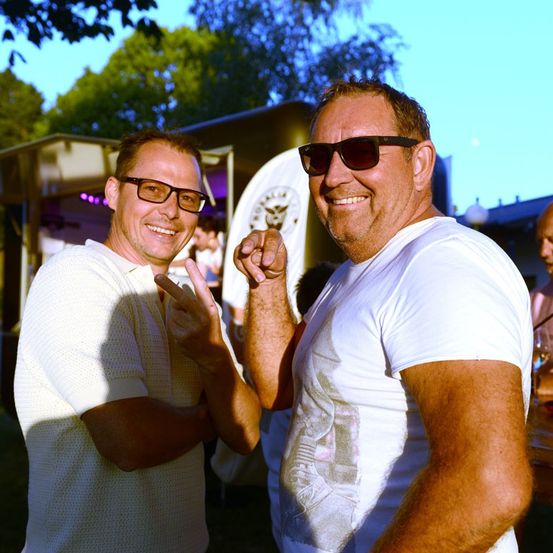 Zwei Männer stehen zusammen und lächeln, einer macht ein Friedenszeichen und der andere trägt eine Sonnenbrille. Sie stehen vor einem Food Truck.