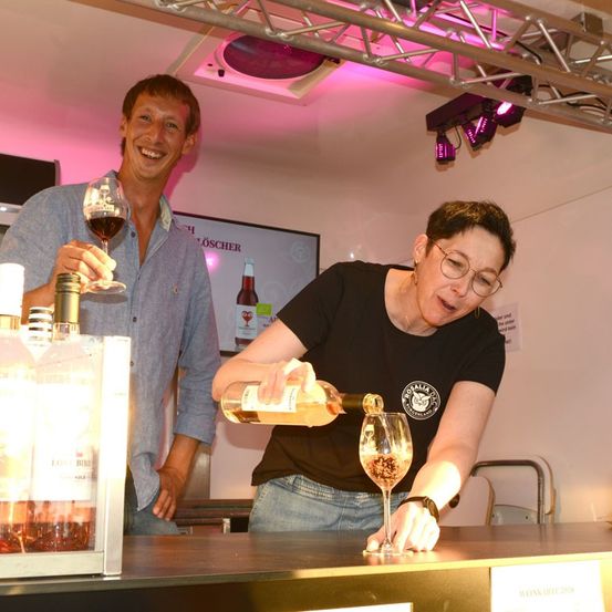 Eine Frau gießt Wein in ein Glas, während ein Mann ein Glas Wein hinter ihr hält. Sie sind in einem Raum mit rosa Licht und einem Monitor an der Wand.