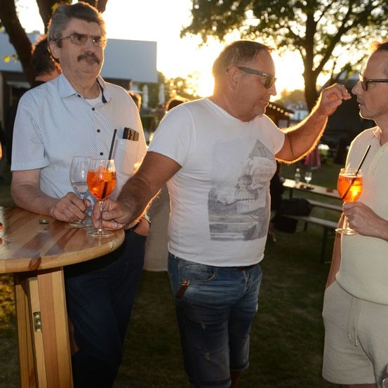 Drei Männer stehen draußen, einer hält ein Glas mit orangefarbenem Getränk, ein anderer gestikuliert und der dritte hält ein Glas mit orangefarbenem Getränk. Sie befinden sich in einem Grasbereich mit einem Holztisch und Bänken.