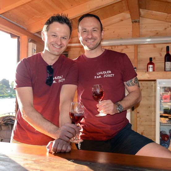 Zwei Männer in burgunderroten T-Shirts, einer hält ein Weinglas, lächeln in einer Holzhütte mit einem Regal mit Flaschen im Hintergrund.