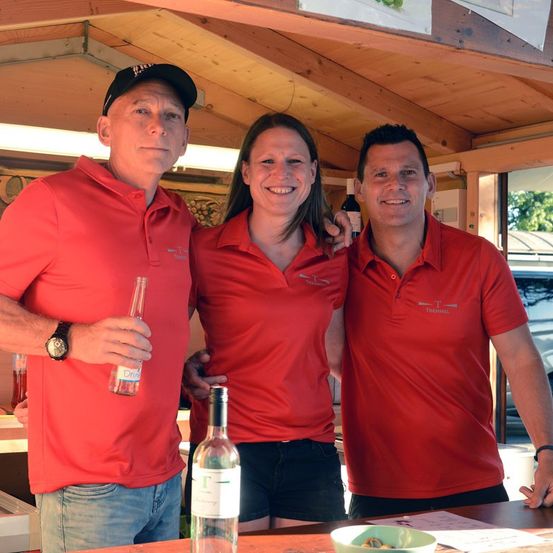 Drei Personen in roten Poloshirts stehen in einer hölzernen Kabine und lächeln für ein Foto. Ein Mann auf der linken Seite hält eine Flasche, eine Frau in der Mitte lächelt und ein Mann auf der rechten Seite hat eine Weinflasche.