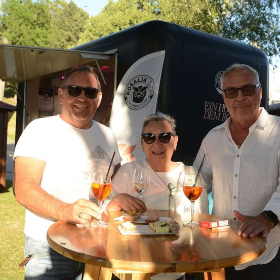 Drei Menschen lächeln und posieren für ein Foto bei einer Outdoor-Veranstaltung. Sie halten Weingläser mit Getränken und vor ihnen steht ein Tisch mit Essen und einer Schachtel Zigaretten. Hinter ihnen befindet sich ein schwarzer Food-Truck mit einem Logo.