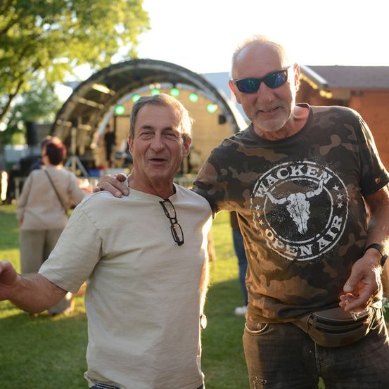 Zwei Männer stehen auf einem Rasenfeld, lächeln und posieren für ein Foto. Einer trägt ein Camouflage-Shirt, der andere ein weißes Shirt. Hinter ihnen steht eine Bühne mit grünen Lichtern und einer Menschenmenge.