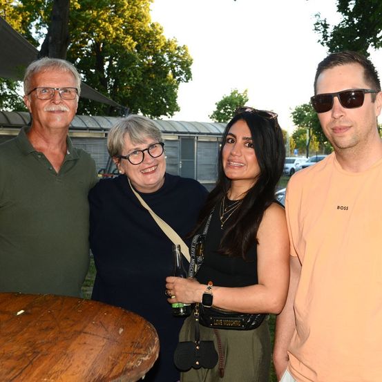 Vier Menschen stehen lächelnd zusammen in einem Park. Ein Mann in Grün hält eine Flasche, eine Frau in Schwarz hält eine Flasche und eine Tasche und eine Frau in Grün hält eine Flasche. Ein Mann in Orange steht rechts.