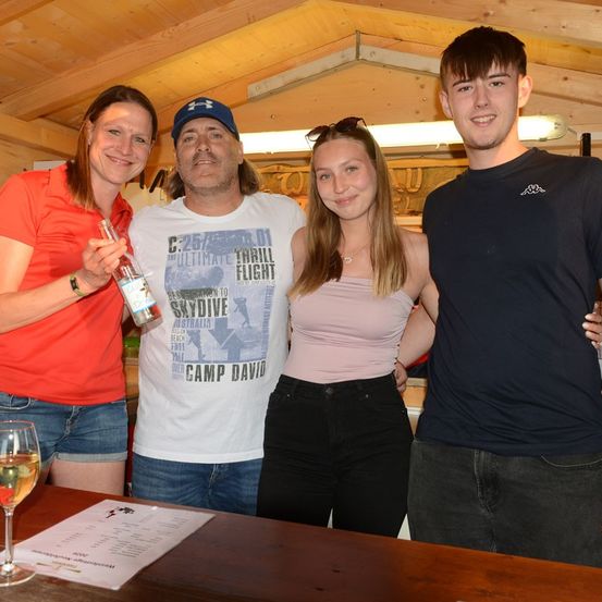 Vier Personen stehen in einem Raum und lächeln für ein Foto. Eine Frau in Rot hält eine Flasche, und ein Mann in Weiß trägt eine Kappe. Eine Frau in Pink steht zwischen zwei Männern. Ein Glas Wein steht auf einem Tisch.