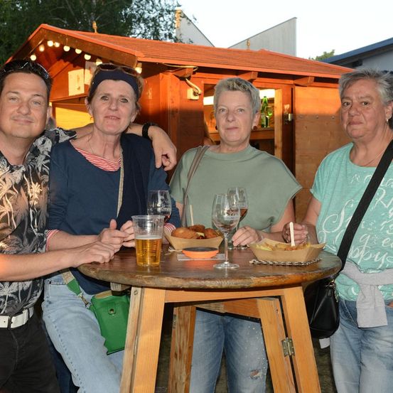 Vier Personen stehen um einen Tisch mit Getränken und Essen und lächeln für ein Foto. Dahinter steht eine kleine Holzstruktur mit Lichtern.
