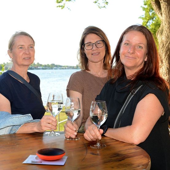 Drei Frauen sitzen an einem Tisch am See, lächeln und halten Weingläser. Die Frau auf der linken Seite trägt ein blaues Shirt, die mittlere trägt eine Brille und ein braunes Shirt, und die rechte trägt ein schwarzes Shirt. Ein kleiner orangefarbener Teller steht auf dem Tisch.