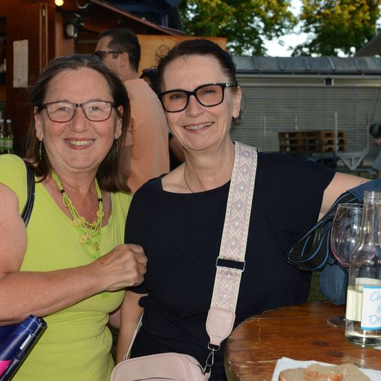 Zwei Frauen mit Brille und Lächeln, eine mit grünem Shirt und die andere mit schwarzem Shirt. Sie sitzen an einem Tisch mit einer Flasche Wein und einem Glas.