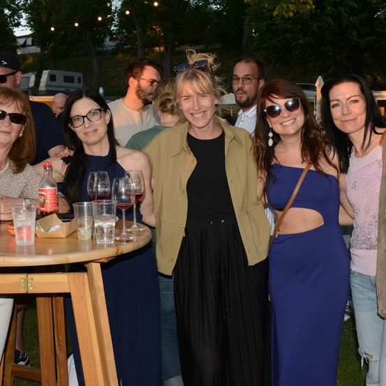 Eine Gruppe von Frauen und Männern versammelt sich bei Nacht im Freien, lächelt und posiert für ein Foto. Der Tisch vor ihnen hat Weingläser, eine Flasche und Tassen. Bäume und ein Gebäude sind im Hintergrund.