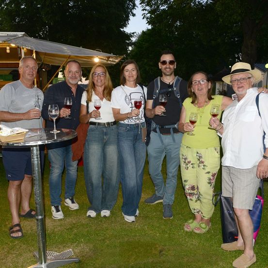 Eine Gruppe von Menschen steht auf einem Rasen und hält Weinglas in der Hand, während sie für ein Foto lächeln. Sie sind lässig gekleidet und scheinen eine gesellige Zusammenkunft zu genießen. Hinter ihnen steht ein Zelt mit einem Tisch und etwas Essen.