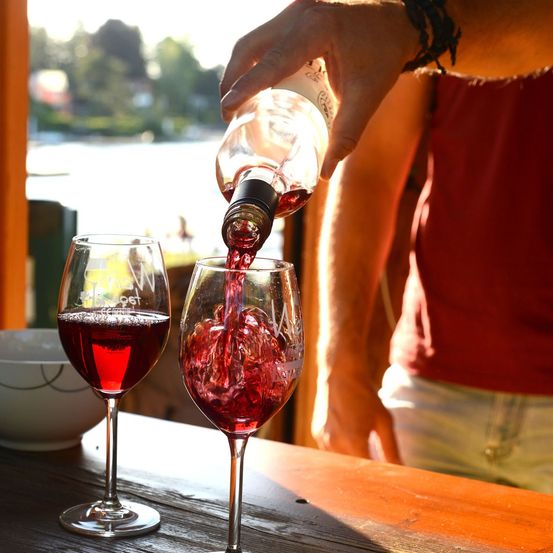 Eine Person gießt roten Wein in zwei Weingläser auf einem Holztisch, mit einer Schüssel in der Nähe.