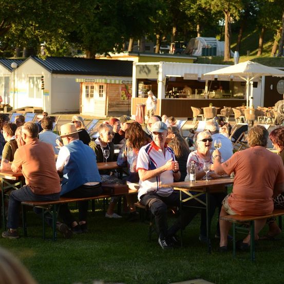 Eine Gruppe von Menschen sitzt auf Bänken um Tische herum in einem Außenbereich, möglicherweise einem Park. Einige halten Weingläser. Im Hintergrund befinden sich Zelte, ein Imbissstand und Bäume.