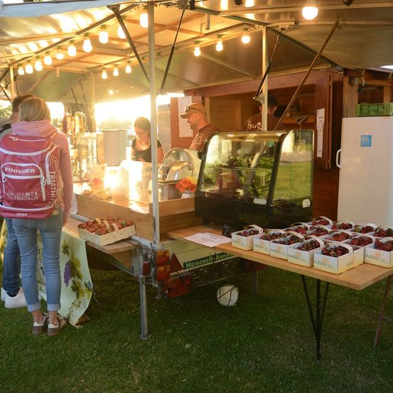 Ein Essensstand mit einem Vordach ist auf einem Rasen aufgestellt. Eine Frau mit einem Rucksack steht vor dem Stand, und ein Mann ist drinnen. Es gibt Erdbeeren in Kisten auf dem Tisch.