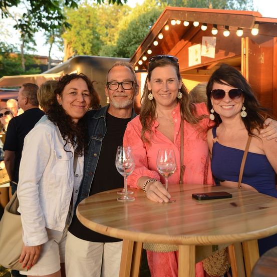 Vier Erwachsene, zwei Frauen und zwei Männer, lächeln und posieren für ein Foto bei einer Outdoor-Veranstaltung. Sie stehen um einen Holztisch mit Weingläsern. Hinter ihnen sind mehrere Leute und ein Food Truck.