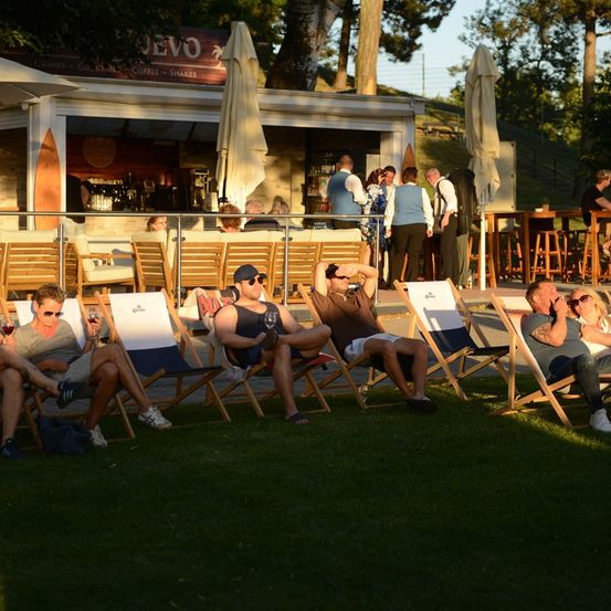 Eine Gruppe von Menschen entspannt sich auf Liegestühlen vor einer Bar, einige mit Getränken in der Hand, im Schatten von Sonnenschirmen.