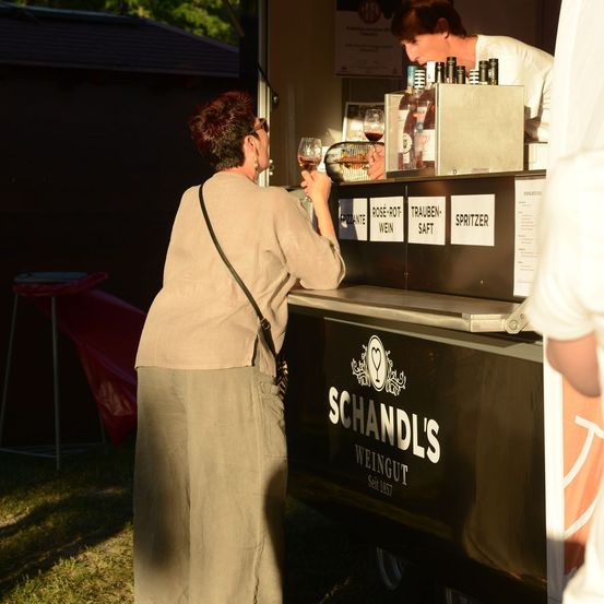 Eine Frau mit einem Weinglas steht vor einem Schandl's Weinstand, mit einem Barkeeper dahinter. Der Tresen hat verschiedene Weinflaschen und ein Menübrett.