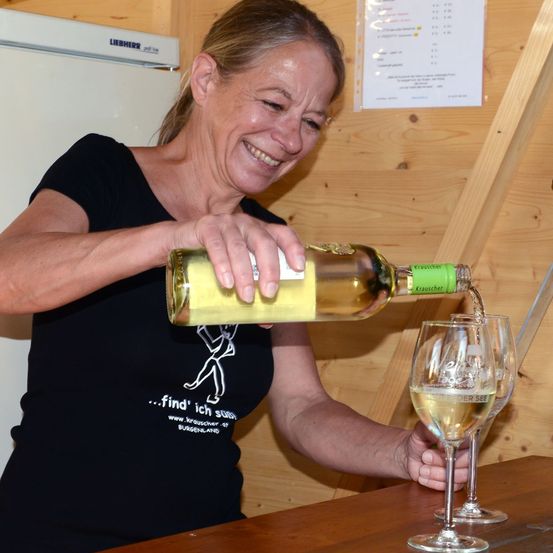 Eine Frau in einem schwarzen T-Shirt schenkt Weißwein in ein Weinglas ein. Sie lächelt und scheint den Moment zu genießen. Die Weinflasche hat ein grünes Etikett.