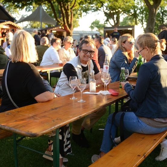 Eine Gruppe von Menschen sitzt an einem langen Holztisch in einer Außenumgebung, möglicherweise einem Biergarten. Sie genießen Getränke und Gespräche. Der Mann in der Mitte trägt eine Brille und lächelt. Auf dem Tisch stehen Flaschen, Gläser und Servietten. Im Hintergrund sitzen und stehen weitere Menschen, und es gibt Bäume und ein Zelt.