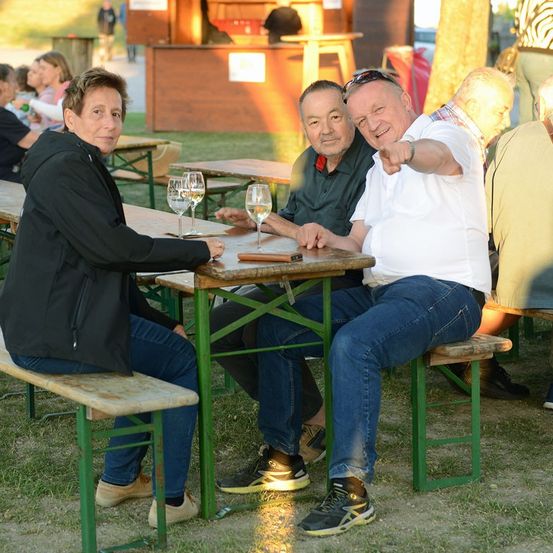 Drei Männer sitzen an einem Tisch in einem Biergarten im Freien, einer zeigt und lächelt, ein anderer mit Weingläsern.