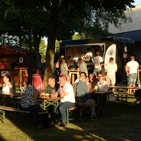 Eine Gruppe von Menschen sitzt um Tische herum in einer Outdoor-Umgebung, möglicherweise bei einem Food-Truck-Event. Einige sitzen, während andere in der Nähe eines Food-Trucks stehen. Bäume und ein Gebäude sind im Hintergrund zu sehen.