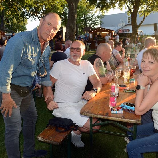 Drei Erwachsene, zwei Männer und eine Frau, sitzen an einem Picknicktisch im Freien, lächeln und schauen in die Kamera. Der Mann auf der linken Seite trägt ein Jeanshemd, der Mann in der Mitte trägt ein weißes T-Shirt und die Frau auf der rechten Seite trägt ein weißes Tanktop.